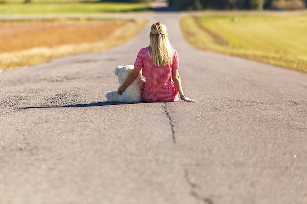 Garota de terno xadrez senta-se na estrada de asfalto com o conceito de ursinho de pelúcia de solidão esperando por felicidade