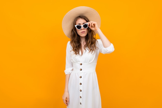 Garota de turista fashionista em um chapéu de palha com óculos escuros, vestido com um vestido branco sobre um fundo amarelo com espaço de cópia