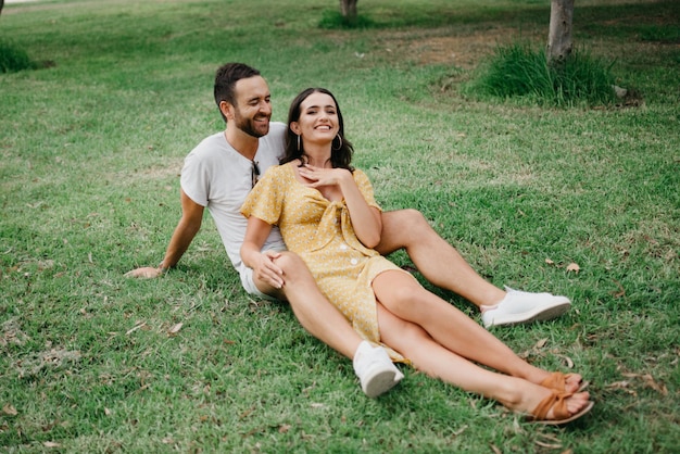 Garota de vestido amarelo está sentada entre as pernas do namorado na grama