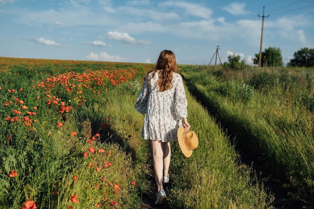 Garota de vestido branco e chapéu de palha caminha entre flores de papoula vermelhas no pôr do sol