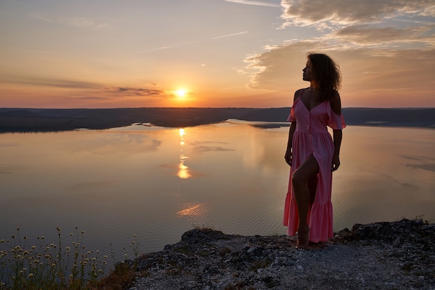 Garota de vestido longo rosa na rocha sobre o lago na noite.