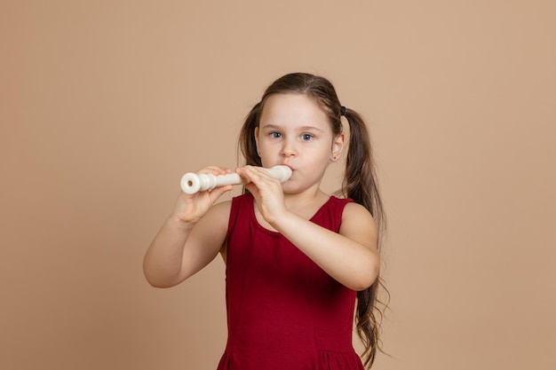 Garota de vestido vermelho pensativamente toca melodia na flauta soprando ar no fundo bege do duto Aprenda a tocar instrumento musical de sopro Flauta e crianças é conceito de desenvolvimento de educação musical