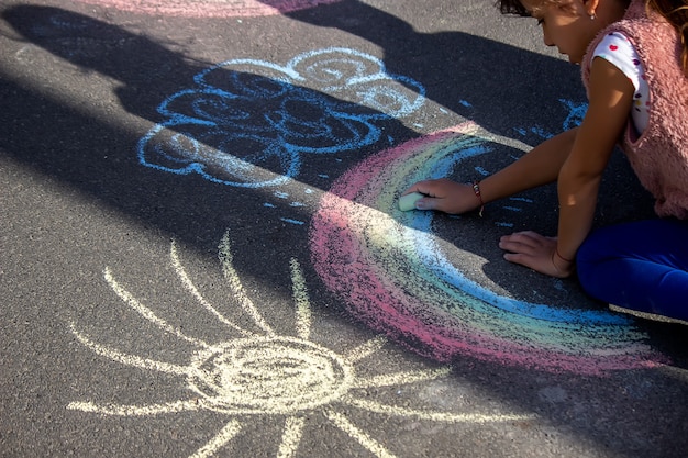 Garota desenha um arco-íris, sol, carro com giz no asfalto. Foco seletivo