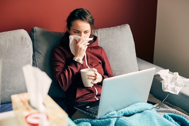 Garota doente trabalha remotamente em casa com uma xícara de chá e lenços na cama, uma freelancer com vírus fica na cama