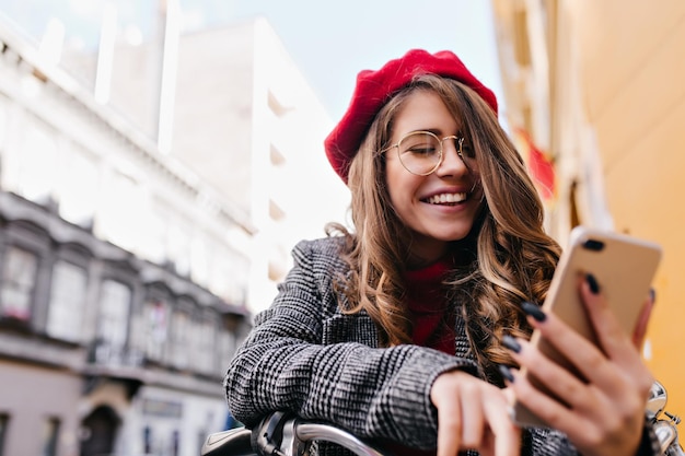 Garota em êxtase em óculos elegantes lê a mensagem de telefone com um sorriso no fundo da cidade de borrão. Retrato ao ar livre de uma jovem muito francesa com o smartphone na mão.