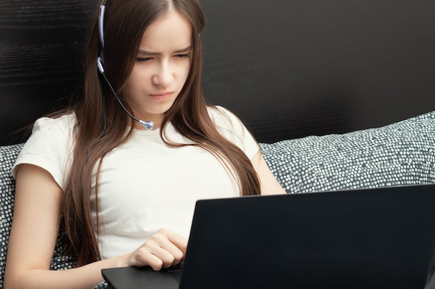 Garota em fones de ouvido com microfone na frente de um laptop está na cama