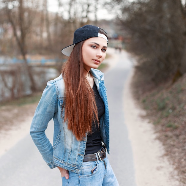 Garota em um boné preto e jaqueta jeans em pé no fundo do parque primavera