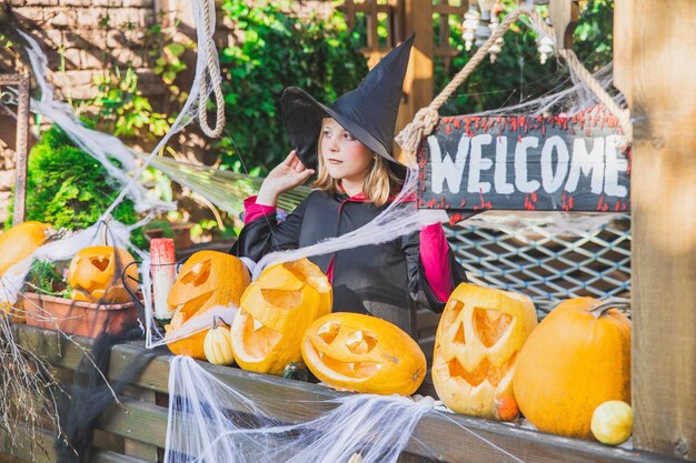 garota em um manto e um chapéu fica perto das abóboras de Halloween