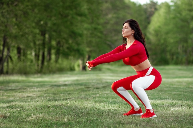 Garota em um terno de esportes faz agachamentos, exercícios físicos em um plano de fundo da natureza. O conceito de um estilo de vida saudável, exercício, ar fresco. Copie o espaço.