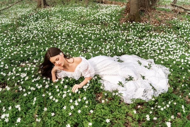 Garota em um vestido de renda branca encontra-se em uma clareira com prímulas Uma jovem de cabelo comprido coleta anêmonas na floresta de primavera