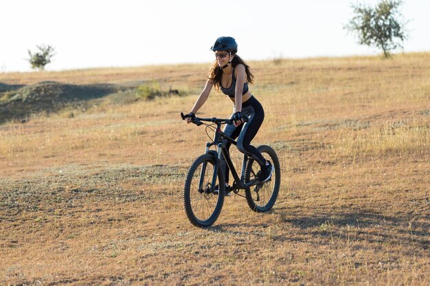 Garota em uma bicicleta de montanha em offroad belo retrato de um ciclista ao pôr do sol Garota fitness monta uma moderna bicicleta de montanha de fibra de carbono em roupas esportivas Closeup retrato de uma garota em um capacete e óculos