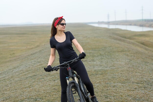 Garota em uma bicicleta de montanha em offroad belo retrato de um ciclista em tempo chuvoso Garota fitness monta uma moderna bicicleta de montanha de fibra de carbono em roupas esportivas Closeup retrato de uma garota de bandana vermelha