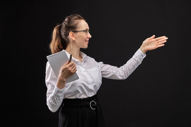Garota em uma camisa branca, segurando um caderno e apontando com a mão em um fundo preto.
