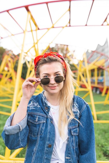 Garota em uma jaqueta jeans e óculos de sol fica no fundo de um parque de diversões, olha para a câmera e sorri