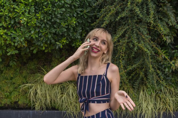 Garota empreendedora em um parque empresarial em uma parede verde de plantas, jovem loira sorrindo fazendo uma ligação com o telefone