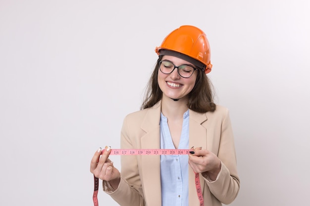 Garota engenheira em um capacete de construção laranja com uma fita métrica nas mãos em um fundo branco