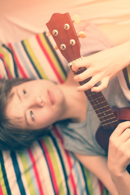 Foto garota engraçada adolescente de pijama cinza deitada na cama com ukulele na mão no travesseiro listrado colorido toned