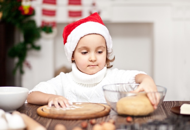 Garota engraçada em um boné de Papai Noel fazendo biscoitos de Natal em casa.