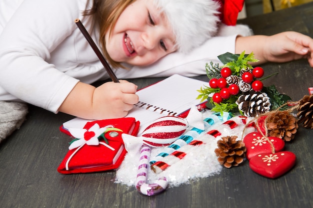 Garota engraçada no chapéu de papai noel escreve carta para o papai noel perto de natal de