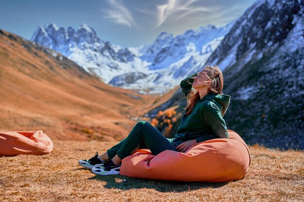 Garota errante sentada em uma cadeira macia durante o descanso em um acampamento. viajante apreciando a paisagem panorâmica enquanto viaja em um vale na montanha