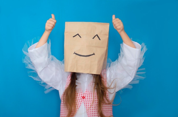 Garota esconde o rosto atrás de um sorriso emoji feliz Foto de uma senhora anônima incógnita segurando um rosto sorridente vestindo uma camisa xadrez contra um fundo azul isolado Ele aponta o dedo para cima