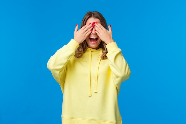 Garota esperando por amigos, diga-lhe os olhos abertos, feche-o com as mãos e conte dez durante a festa de aniversário, namorada traga presente surpresa, sorrindo com emoção, jogando peekaboo, fique em uma parede azul