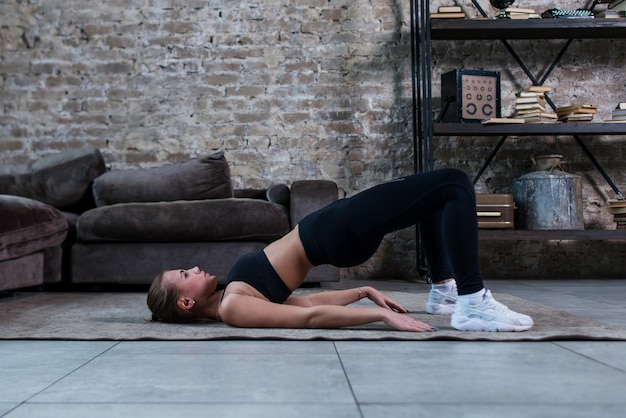 Garota esportiva fazendo levantamento de quadril no chão ou exercício de elevação de bunda deitada no chão em seu apartamento loft