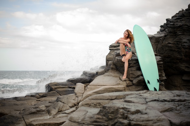 Garota esportiva no maiô multicolor, sentada perto da prancha de surfe na rocha sobre o oceano Atlântico e céu claro