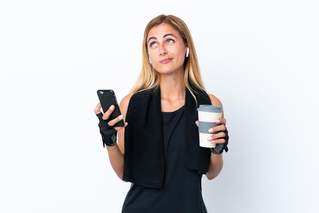 Garota esportiva uruguaia loira isolada em fundo branco segurando café para levar e um celular enquanto pensava em algo