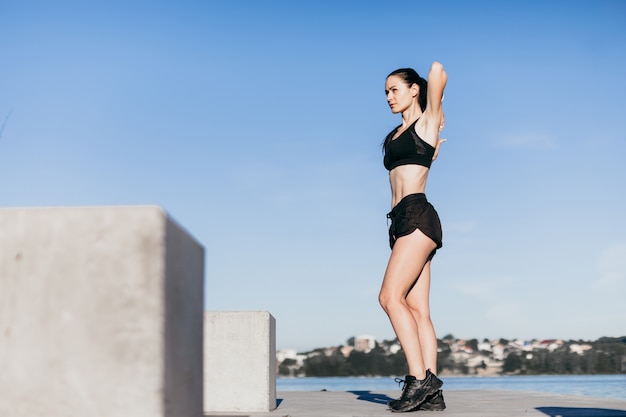 Garota esportiva, vestida de preto, aquece antes de correr, no playground da praia da cidade de manhã