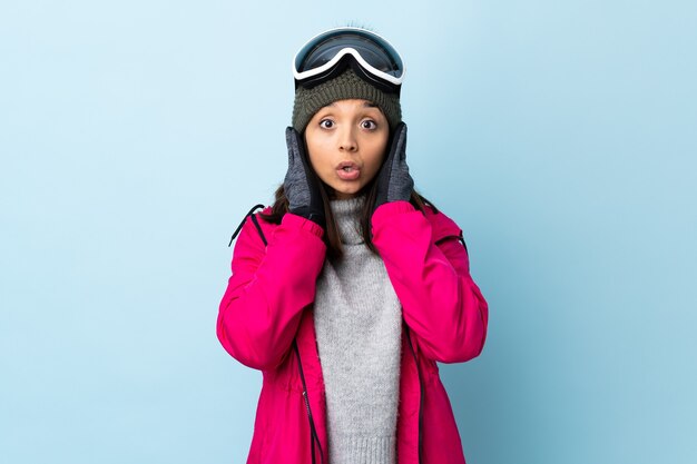 Garota esquiadora de raça mista com óculos de snowboard sobre fundo azul isolado frustrada e cobrindo as orelhas.