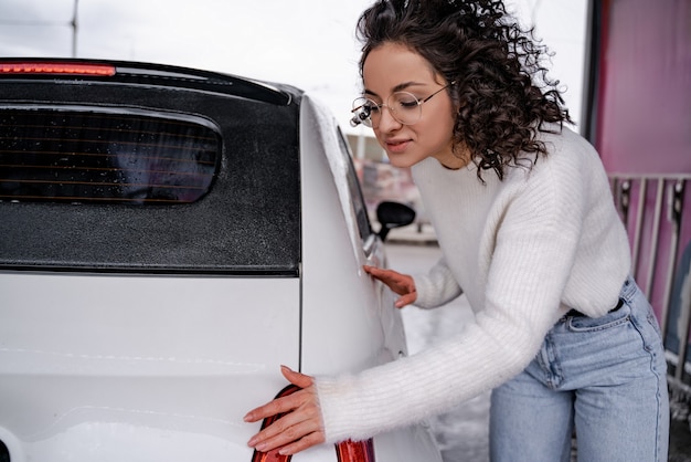 Garota europeia assistindo automóvel pessoal após a lavagem na lavagem de carros self-service. Mulher jovem e sorridente, concentrada, usando óculos