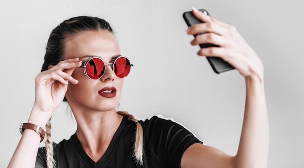 Garota fashion de óculos escuros e óculos coloridos fazendo selfie no telefone