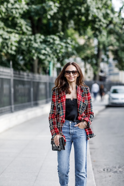 Garota fashion em uma jaqueta xadrez e jeans andando pela cidade com uma bolsa preta.