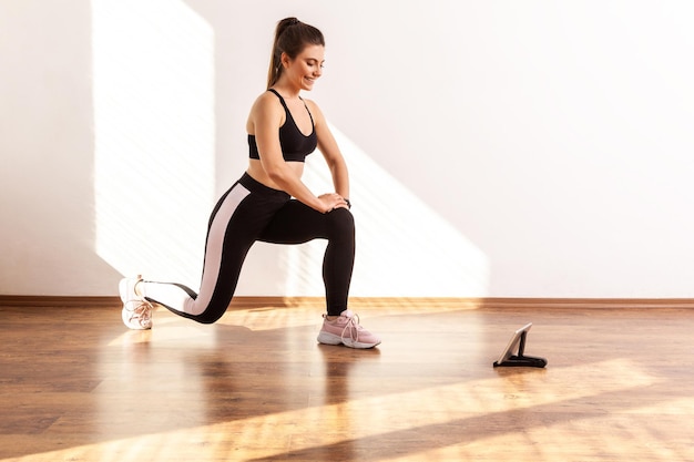 Foto garota fazendo agachamento ou agachamento dividido, repetindo após o blogueiro de fitness, assistindo a um vídeo de treinamento no tablet, vestindo top esportivo preto e meia-calça. tiro de estúdio de corpo inteiro iluminado pela luz do sol da janela.