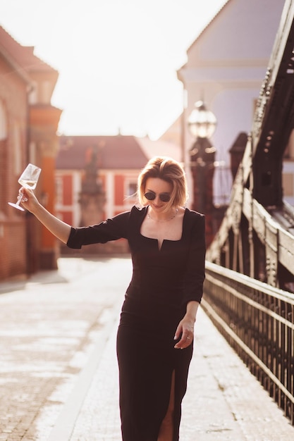 Garota feliz atraente em um vestido preto caminha por uma rua da cidade com um copo de vinho Estilo de vida glamoroso Moda