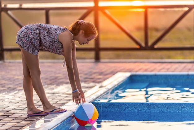 Garota feliz bronzeada gorda com cabelo escuro trançado em coque em terno de verão brilhante está jogando perto da piscina com bola inflável no contexto do sol dourado da noite de verão