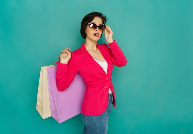Garota feliz caucasiana com sacolas de compras e óculos escuros. Compradora viciada em compras no fundo do estúdio turquesa com espaço de cópia