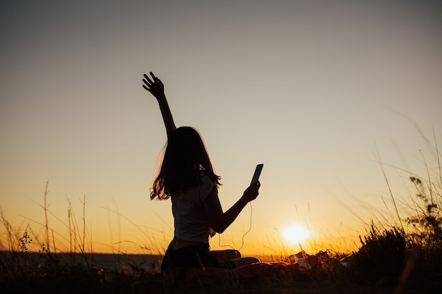 Garota feliz com a mão cantando e ouvindo música em fones de ouvido em um campo ao pôr do sol incrível.