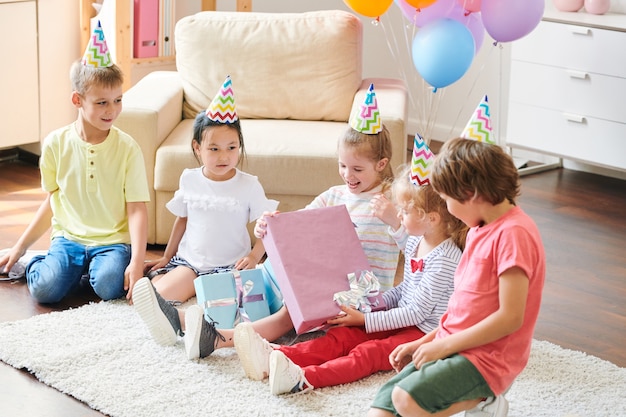 Garota feliz com boné de aniversário desempacotando uma grande caixa de presente enquanto está sentada no chão entre seus amigos em uma festa em casa