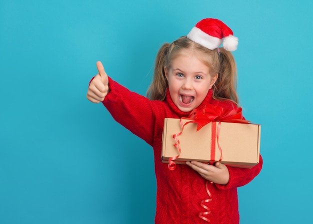 Garota feliz com presentes no chapéu de Papai Noel mostrando o polegar para cima gesto