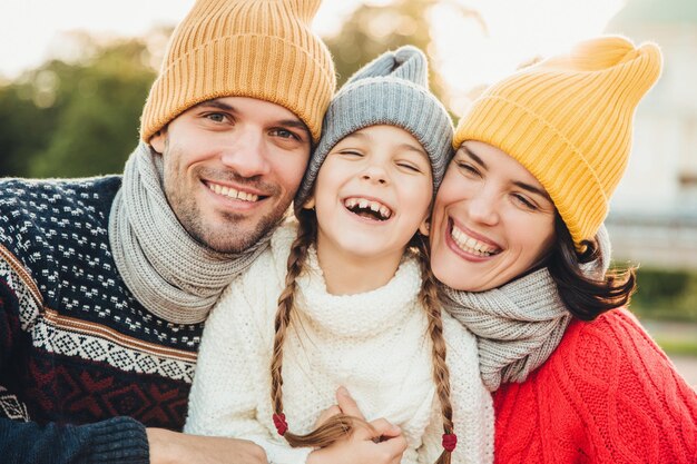 Garota feliz com tranças usa suéter de malha quente fica entre pai e mãe riem alegremente têm sorrisos sinceros em seus rostos Família relaxada tem feriados passam bons momentos juntos