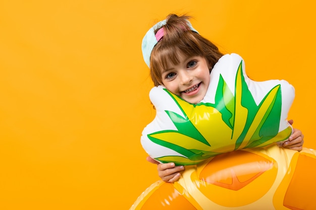 garota feliz com um boné de beisebol em um maiô com um anel de borracha de abacaxi em uma parede laranja