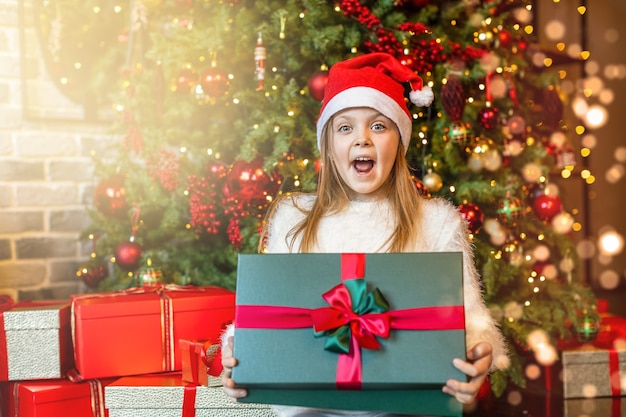 Garota feliz com um chapéu de Papai Noel é surpreendida por uma caixa de presente mágica no fundo da árvore de Natal. Feliz Natal