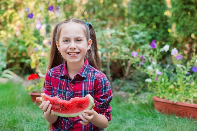 Garota feliz comendo melancia no jardim. as crianças comem frutas ao ar livre. lanche saudável para crianças. menina brincando no jardim, mordendo uma fatia de melancia.