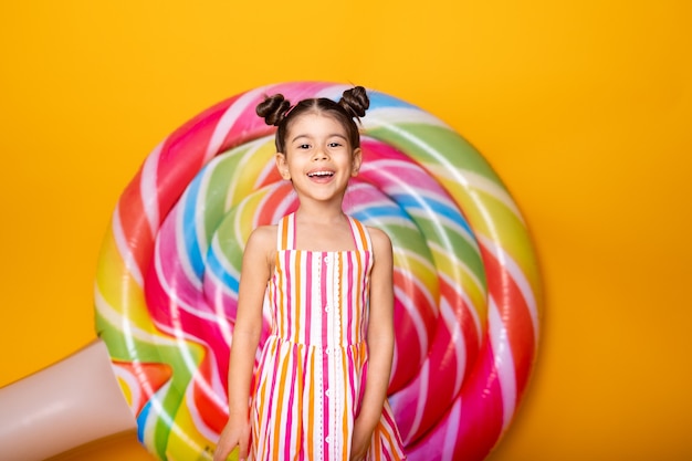 Garota feliz e animada criança árabe em vestido listrado colorido se divertindo na superfície amarela com pirulito