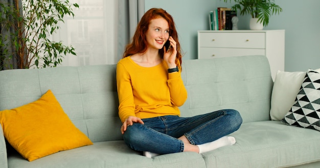 Garota feliz e positiva, sentado no sofá em casa e falando em seu smartphone