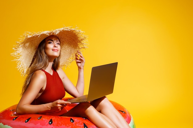 Garota feliz em palha sentada em um anel inflável trabalhando em um laptop em um fundo amarelo.