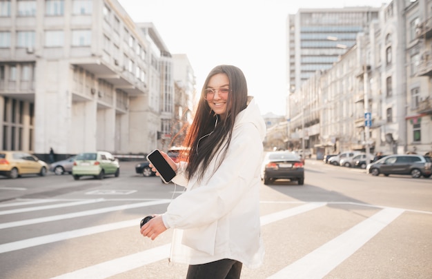 Garota feliz em roupas casuais e óculos de sol com um smartphone na mão, desce a rua contra a cidade durante o sol, olha a câmera e sorri