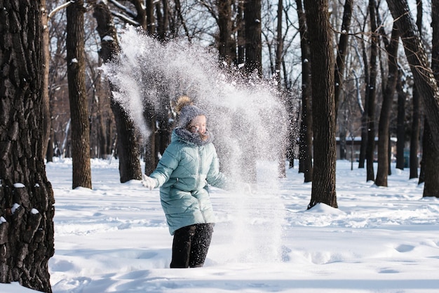 Garota feliz em roupas quentes joga neve na floresta de inverno Caminhe ao ar livre